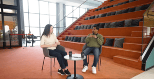 Two people talking in a university space, seated in armchairs in front of wooden amphitheatre-style steps.
