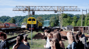 A group of young people observes a yellow railway maintenance locomotive in an industrial railway area.
